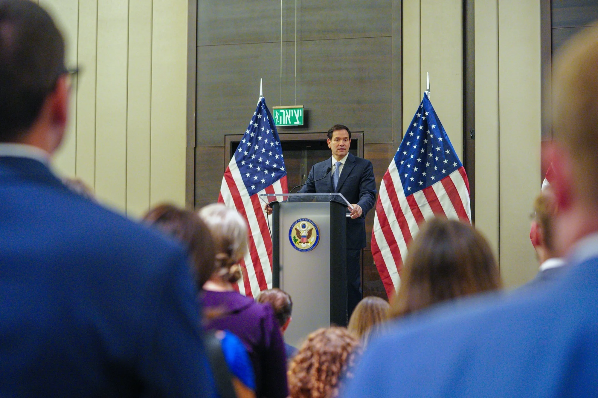 Marco Rubio standing at a podium, speaking with U.S. flags on both sides and a few people seated in the audience.