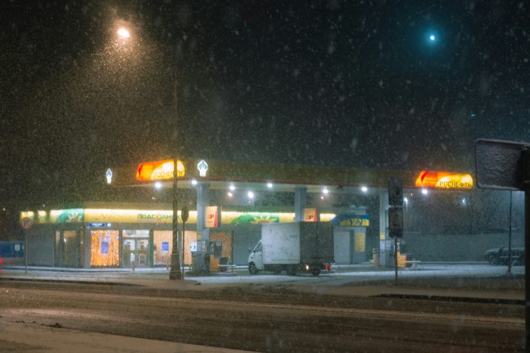 A small gas station glows under harsh white lights at night, its pumps partially buried in drifting snow. Thick flakes fall diagonally across the scene, blurring the dark sky and coating the ground in a fresh layer of white. The surrounding area is quiet and empty, giving the station an isolated, weather-beaten look in the middle of the snowstorm.